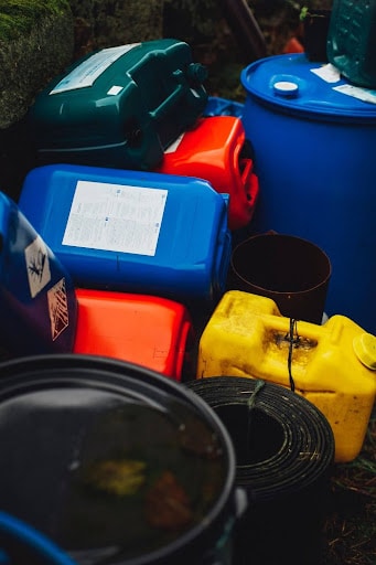 A variety of colorful industrial containers for hazardous waste management, including blue, red, and yellow drums and jugs, some with hazard labels, properly stored for environmental safety compliance.